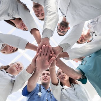 Eine Gruppe von medizinischem Personal in weißen Kitteln legt ihre Hände übereinander in der Mitte, symbolisiert Teamarbeit und Zusammenarbeit.