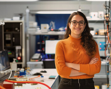Eine Frau mit Brille steht in einem technischen Labor, umgeben von Geräten und Kabeln. Sie trägt einen orangefarbenen Pullover und hat die Arme verschränkt. Im Hintergrund sind Computer und technische Ausrüstung sichtbar.