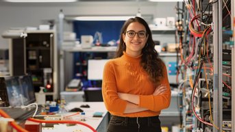 Eine Frau mit Brille steht in einem technischen Labor, umgeben von Geräten und Kabeln. Sie trägt einen orangefarbenen Pullover und hat die Arme verschränkt. Im Hintergrund sind Computer und technische Ausrüstung sichtbar.