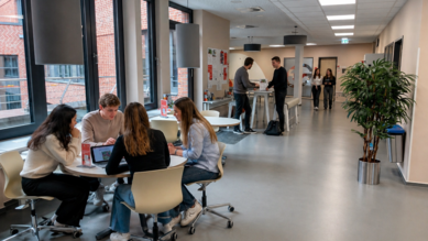 Studierende arbeiten an Tischen in einem modernen Raum mit großen Fenstern. Einige Personen stehen und unterhalten sich im Hintergrund.