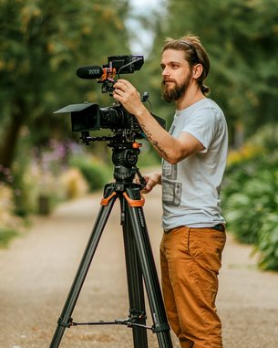 Ein Mann mit Bart steht neben einem Stativ, auf dem eine Kamera montiert ist. Er justiert das Mikrofon und die Kamera, während er in einem Garten mit grünen Pflanzen und Blumen arbeitet.