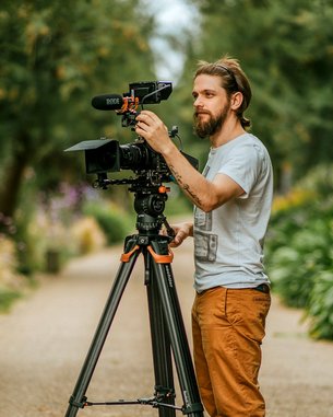 Ein Mann mit Bart steht neben einem Stativ, auf dem eine Kamera montiert ist. Er justiert das Mikrofon und die Kamera, während er in einem Garten mit grünen Pflanzen und Blumen arbeitet.