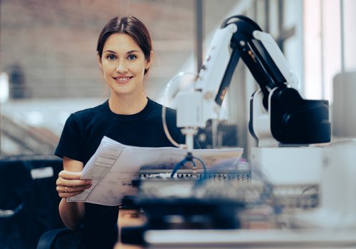Eine Frau hält Dokumente in der Hand und lächelt, während ein Roboterarm neben ihr steht. Sie befindet sich in einer modernen Arbeitsumgebung.