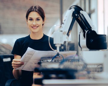 Eine Frau hält Dokumente in der Hand und lächelt, während ein Roboterarm neben ihr steht. Sie befindet sich in einer modernen Arbeitsumgebung.