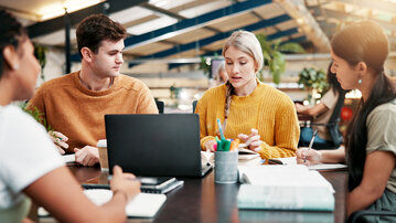 Fünf Personen sitzen an einem Tisch, diskutieren und arbeiten an Laptops und Notizen in einem modernen, hellen Raum.