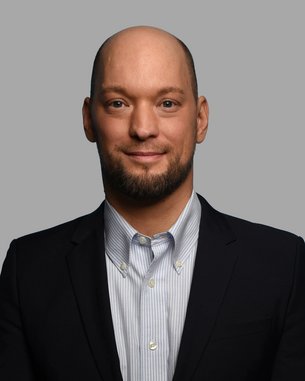 Ein Mann mit Glatze und Bart trägt einen Anzug und ein Hemd mit Streifen. Er blickt direkt in die Kamera.
