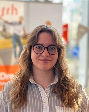 Junge Frau mit lockigem, blondem Haar und Brille, trägt ein gestreiftes Hemd. Im Hintergrund sind unscharfe Banner und Fenster zu sehen.