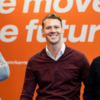 Vier Männer stehen und sitzen in einem modernen Büro, lächelnd vor einer orangefarbenen Wand mit dem Text „We move the future“.