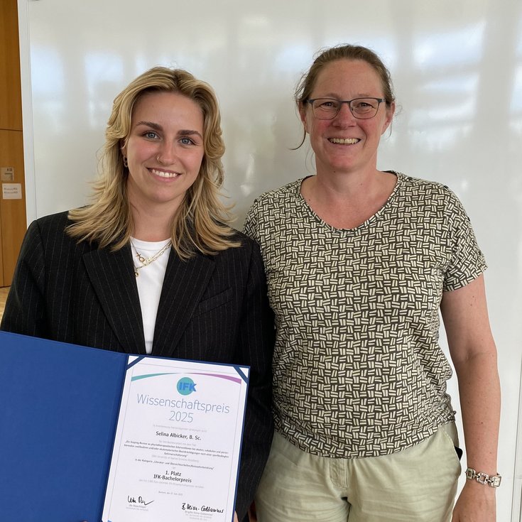 Zwei Frauen stehen nebeneinander, eine hält ein Zertifikat für den Wissenschaftspreis 2025 in der Hand. Beide lächeln in die Kamera.