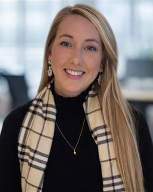 Eine Frau mit langen, blonden Haaren trägt eine schwarze Bluse und einen karierten Schal. Sie lächelt und steht in einem modernen Büro.