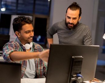 Two men are working at a computer in a modern office. One is pointing at the screen while the other watches attentively.