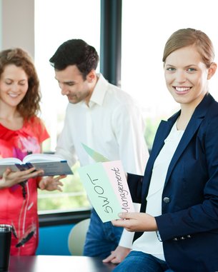 Drei Personen in einem modernen Büro, eine Frau hält ein Blatt mit der Aufschrift "SWOT Management", während die anderen beiden ein Buch betrachten.