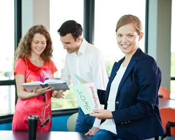 Drei Personen in einem modernen Büro, eine Frau hält ein Blatt mit der Aufschrift "SWOT Management", während die anderen beiden ein Buch betrachten.