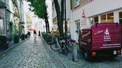 Eine gepflasterte Straße mit einem lila Lieferwagen der Bremer Bonbonfabrik und Fahrrädern, umgeben von Bäumen und Gebäuden.