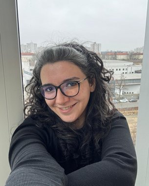Eine Person mit lockigem, schwarzem Haar und Brille lächelt vor einem Fenster mit städtischem Hintergrund.