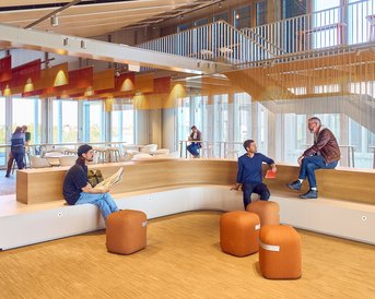 Moderne Innenarchitektur mit Sitzgelegenheiten, mehreren Personen, die sich entspannen oder arbeiten, und großen Fenstern, die Tageslicht hereinlassen.