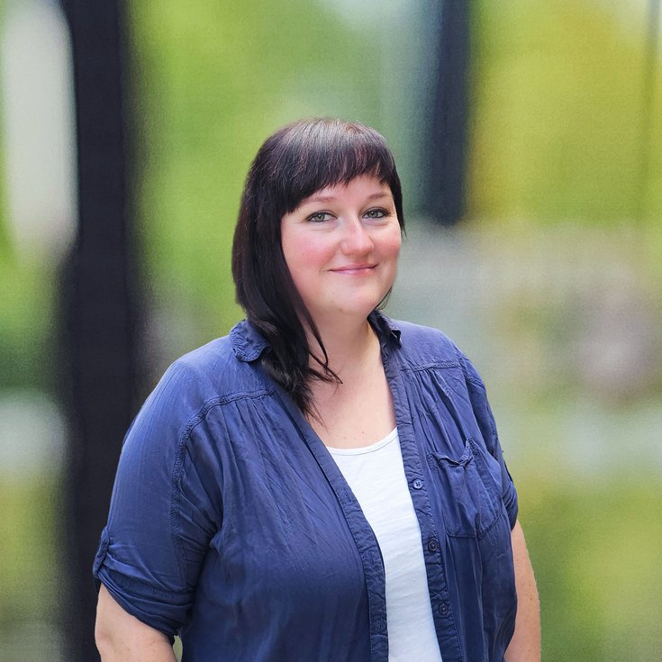 Eine Frau mit dunklen Haaren trägt ein blaues Hemd und steht lächelnd vor einem unscharfen, grünen Hintergrund.