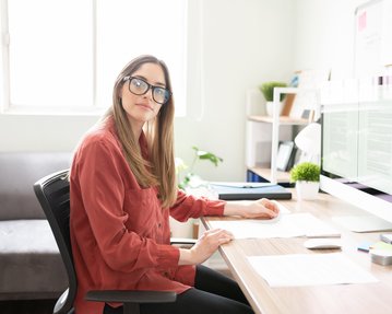 A woman with glasses is sitting at a desk, looking at the camera while working on a computer. Papers and office supplies are scattered on the table.