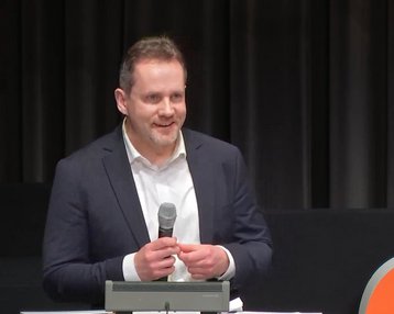 Ein Mann in einem Anzug hält ein Mikrofon und spricht auf einer Bühne. Im Hintergrund sind Vorhänge und ein orangefarbenes Logo sichtbar.