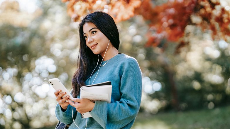 Eine junge Frau mit langen, dunklen Haaren steht im Freien. Sie trägt einen blauen Pullover, hört Musik über Kopfhörer und hält ein Smartphone sowie Notizblätter in der Hand. Bunte Herbstblätter sind im Hintergrund sichtbar.