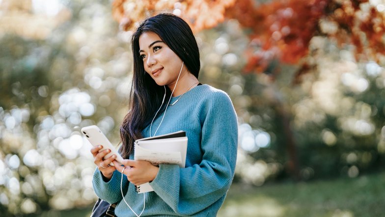 Eine junge Frau mit langen, dunklen Haaren steht im Freien. Sie trägt einen blauen Pullover, hört Musik über Kopfhörer und hält ein Smartphone sowie Notizblätter in der Hand. Bunte Herbstblätter sind im Hintergrund sichtbar.