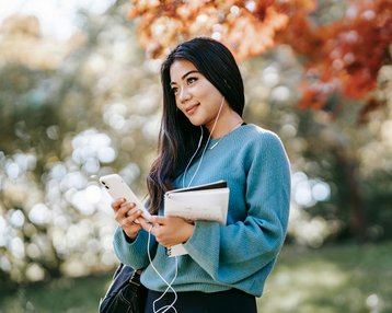 Eine junge Frau mit langen, dunklen Haaren steht im Freien. Sie trägt einen blauen Pullover, hört Musik über Kopfhörer und hält ein Smartphone sowie Notizblätter in der Hand. Bunte Herbstblätter sind im Hintergrund sichtbar.