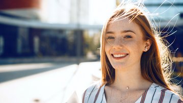 Young woman with long blonde hair and freckles, smiling as she holds a laptop under her arm, standing in an urban setting.