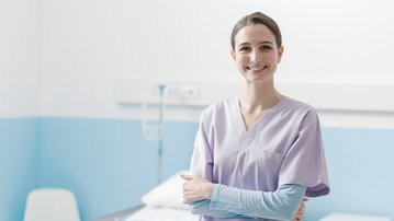 Eine Krankenschwester in lila Uniform steht lächelnd in einem Krankenhauszimmer neben einem Bett.