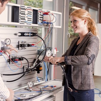 Two people are working on a technical device connected with hoses and measuring instruments. The woman is smiling as she handles a cable, while the man watches attentively.