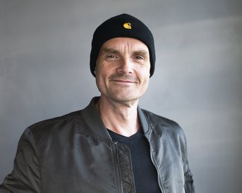 Mann mit lässiger Jacke und schwarzer Mütze steht lächelnd vor einem neutralen Hintergrund.