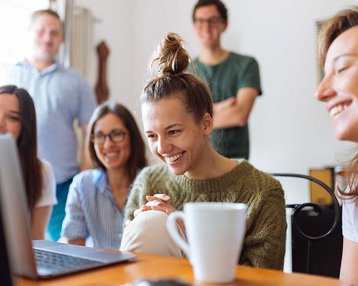 Six people are sitting around a table, looking at a laptop. A woman is smiling while the others watch attentively.