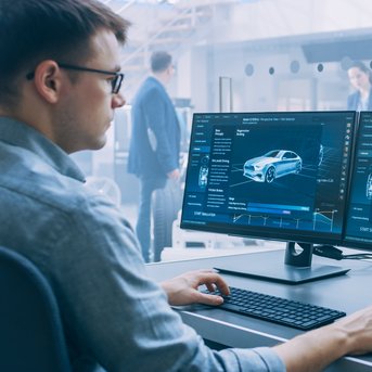 Ein Mann arbeitet an einem Schreibtisch mit zwei Monitoren, die technische Informationen über Autos anzeigen.