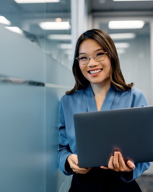 A woman with glasses is holding a laptop in a modern office and smiling warmly. She is wearing a blue blouse.