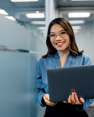 A woman with glasses is holding a laptop in a modern office and smiling warmly. She is wearing a blue blouse.