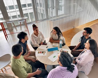 Sechs Personen sitzen um einen Tisch und diskutieren. Auf dem Tisch liegen Unterlagen und Getränke. Der Raum hat große Fenster und modernes Design.