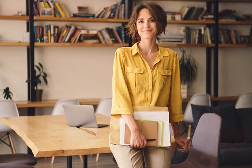 Eine Frau mit lockigem Haar trägt ein gelbes Hemd und steht in einem modernen Raum. Sie hält Notizblöcke und einen Laptop in der Hand. Im Hintergrund sind Bücherregale und ein Tisch mit Stühlen sichtbar.
