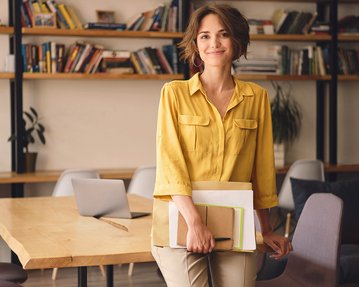 Eine Frau mit lockigem Haar trägt ein gelbes Hemd und steht in einem modernen Raum. Sie hält Notizblöcke und einen Laptop in der Hand. Im Hintergrund sind Bücherregale und ein Tisch mit Stühlen sichtbar.