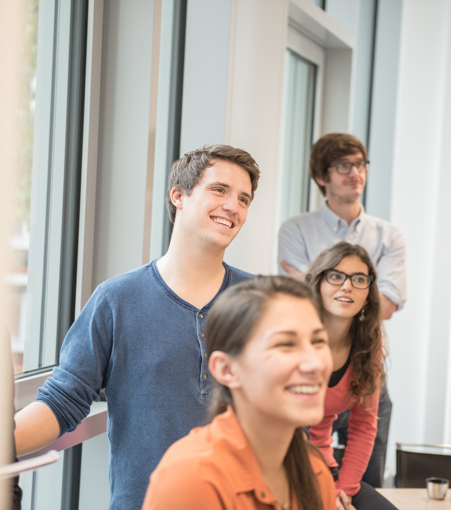 A group of five young adults stands in a bright room. They are smiling and looking attentively in one direction. Two of them are wearing glasses, while the others are dressed casually.
