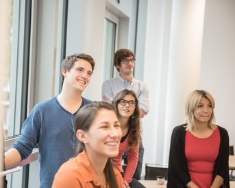 A group of five young adults stands in a bright room. They are smiling and looking attentively in one direction. Two of them are wearing glasses, while the others are dressed casually.