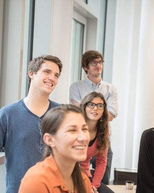 A group of five young adults stands in a bright room. They are smiling and looking attentively in one direction. Two of them are wearing glasses, while the others are dressed casually.