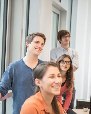 Eine Gruppe von fünf jungen Erwachsenen steht in einem hellen Raum. Sie lächeln und schauen aufmerksam in eine Richtung. Zwei Personen tragen Brillen, während die anderen lässige Kleidung tragen.