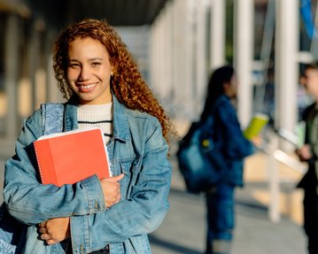 Eine junge Frau mit lockigem, braunem Haar trägt eine Jeansjacke und hält ein rotes Notizbuch. Im Hintergrund sind weitere Studierende zu sehen.