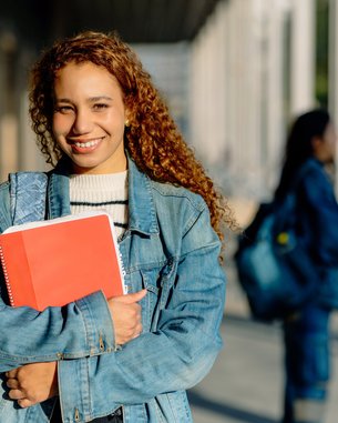 Eine junge Frau mit lockigem, braunem Haar trägt eine Jeansjacke und hält ein rotes Notizbuch. Im Hintergrund sind weitere Studierende zu sehen.