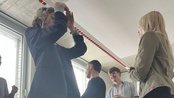 Eine Gruppe von Menschen steht in einem Raum, einige tanzen oder interagieren, während andere zuschauen. Fenster im Hintergrund.