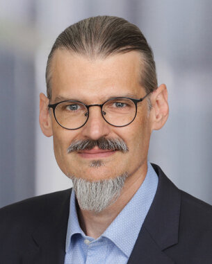 A man with glasses and a beard is wearing a suit and looking directly at the camera. The background is blurred and neutral.