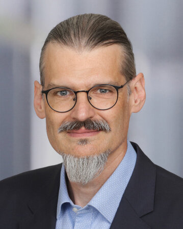 A man with glasses and a beard is wearing a suit and looking directly at the camera. The background is blurred and neutral.