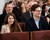 Eine Gruppe von Menschen sitzt in einem Saal, lächelnd und in formeller Kleidung, während sie auf eine Veranstaltung warten.