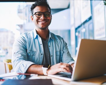 Ein junger Mann mit Brille sitzt an einem Tisch in einem Café und arbeitet an einem Laptop. Er lächelt freundlich.