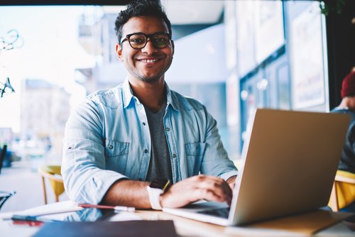 Ein junger Mann mit Brille sitzt an einem Tisch in einem Café und arbeitet an einem Laptop. Er lächelt freundlich.