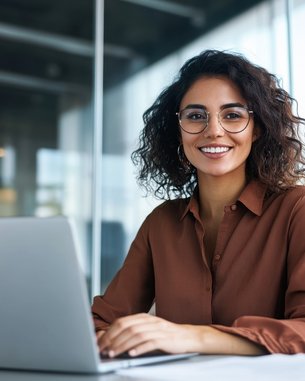 Eine Frau mit lockigem Haar und Brille sitzt an einem Schreibtisch und lächelt, während sie an einem Laptop arbeitet.